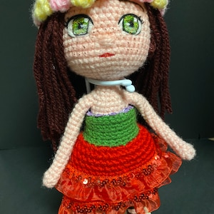 May include: Crocheted doll with brown hair, green eyes, and a pink, yellow, and pink flower crown. The doll is wearing a red dress with green trim and a red, sequined skirt.
