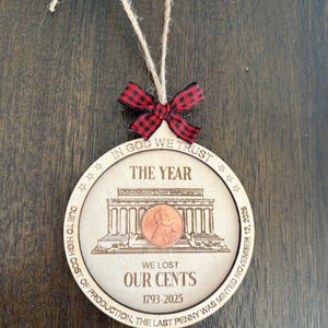 May include: A round wooden Christmas ornament with a red and black plaid bow and twine for hanging. The ornament features engraved text and a penny image. Text includes "IN GOD WE TRUST", "THE YEAR", "WE LOST OUR CENTS", and "1793-2025".