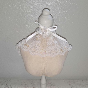 Può includere: Un busto color crema con un colletto in pizzo bianco, ornato di perle e un fiocco di raso. Il busto è montato su un supporto bianco, che mostra un design delicato ed elegante.