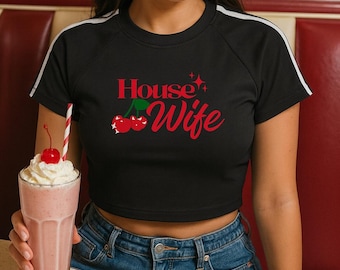 Puede incluir: Top corto negro con rayas blancas en las mangas y las palabras "House Wife" en rojo con gráficos de cerezas. La persona lleva vaqueros azules y sostiene un batido rosa con crema batida y una cereza.