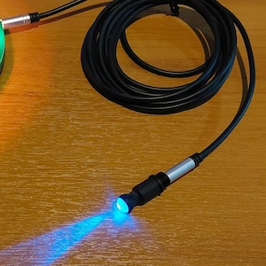 以下が含まれることがあります： 先端に青いLEDライトが付いた黒いケーブル。ケーブルは巻き上げられており、光は木製の表面に当たっています。
