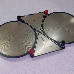 Può includere: Un circuito elettronico nero e oro con due grandi sezioni circolari e sezioni triangolari. La scheda ha terminali neri e rossi. Le sezioni dorate hanno un design a spirale.