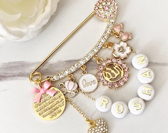 May include: A gold safety pin with a white and pink enamel charm with the word "love" on it. The pin is decorated with rhinestones and has several other charms, including a heart, a carriage, a bow, a flower, and a round charm with Arabic calligraphy. The pin also has white beads with the name "ROSHAN" spelled out.