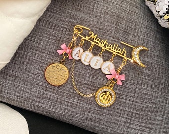 May include: Gold-tone baby pin with a crescent moon charm, a charm with the word "Mashallah" and charms with the letters "ALIA". The pin also has a charm with an Islamic symbol and a charm with Arabic writing.