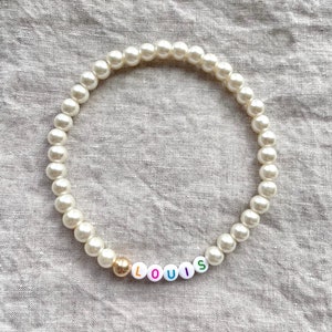 May include: A white pearl necklace with a gold bead and colorful letter beads spelling "LOUIS".