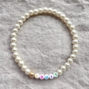 May include: A white pearl necklace with a gold bead and colorful letter beads spelling "LOUIS".