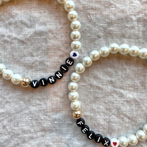 May include: Two white pearl bracelets with black letter beads spelling out the names "Vinnie" and "Felix". Each bracelet has a small heart charm.
