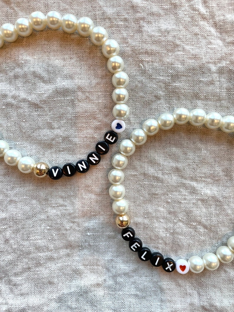 May include: Two white pearl bracelets with black letter beads spelling out the names "Vinnie" and "Felix". Each bracelet has a small heart charm.