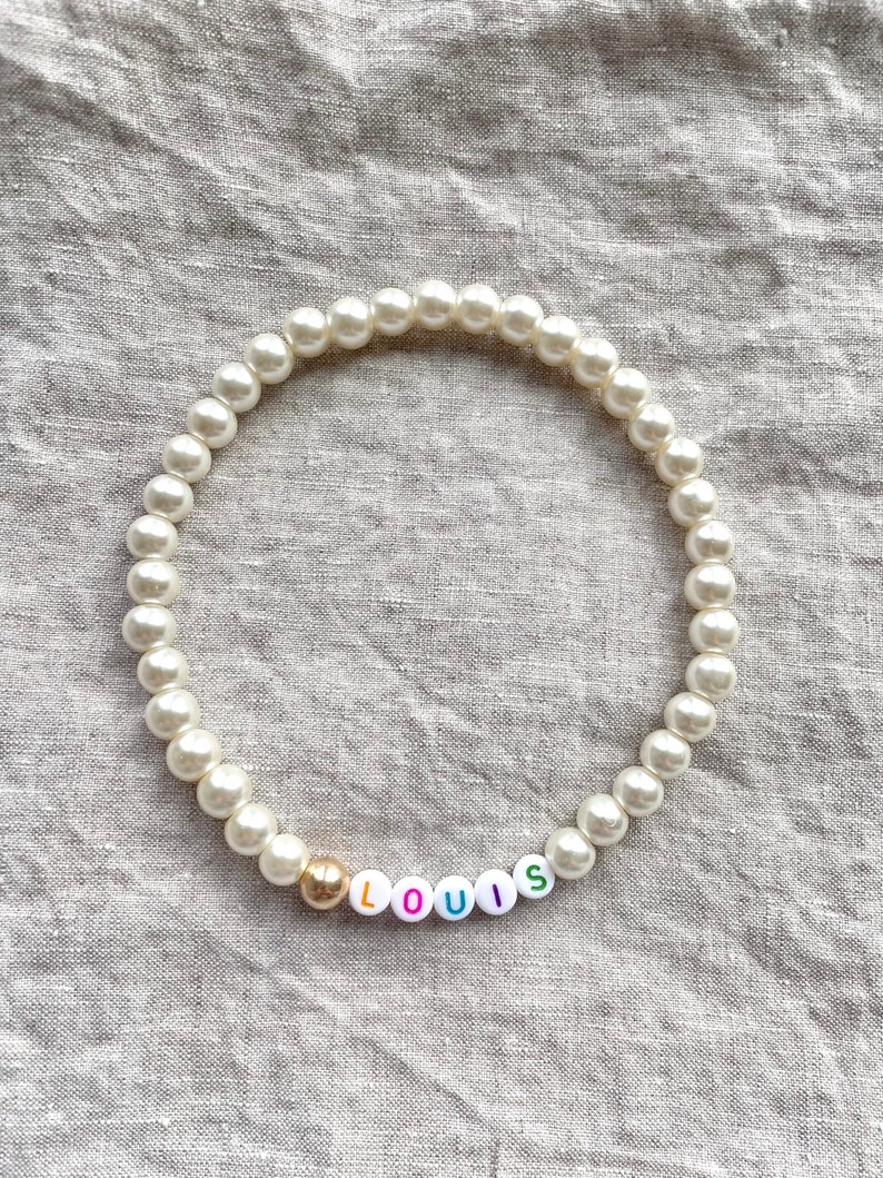 May include: A white pearl necklace with a gold bead and colorful letter beads spelling "LOUIS".