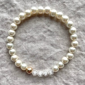 May include: A white pearl bracelet with a gold bead and clear letter beads spelling "BABE".