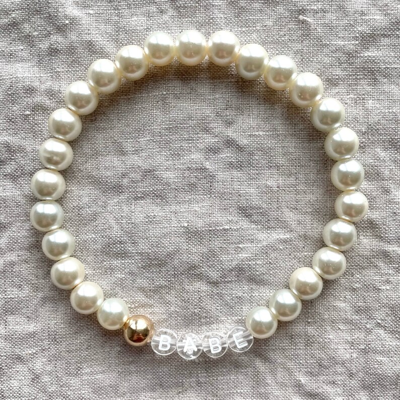 May include: A white pearl bracelet with a gold bead and clear letter beads spelling "BABE".