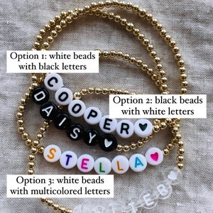 May include: Four different options for letter bead bracelets. The first option has white beads with black letters, the second has black beads with white letters, the third has white beads with multicolored letters, and the fourth has clear beads with white letters. The bracelets spell out the names "COOPER", "DAISY", "STELLA", and "WALTER".