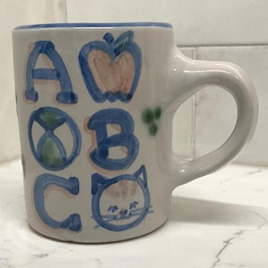 May include: A white ceramic mug featuring hand-painted blue letters A, B, and C. The design includes a blue apple illustration next to the letter A, a circular globe-like graphic next to the letter B, and a blue cat face illustration next to the letter C.