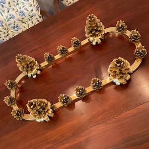 May include: A gold-toned metal candle holder shaped like a pine cone branch. The holder has 10 candle holders, each shaped like a pine cone. The holder is on a wooden table.