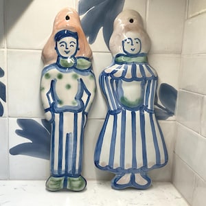 May include: Two vertical ceramic wall plaques shaped like figures. The left figure wears a white top with green dots and blue striped pants. The right figure wears a blue and white striped dress with a ruffled collar. Both have painted faces.