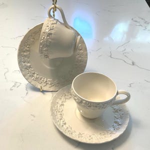 May include: Two off-white teacups and saucers with floral relief patterns. One cup is suspended above its saucer, while the other sits on a table. The saucers and cups have a matching design, creating a cohesive tea set.