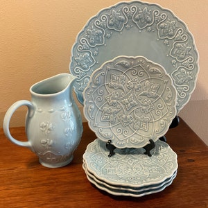 May include: A set of light blue ceramic dinnerware with an intricate embossed floral design. The set includes a pitcher, a large plate, a smaller plate, and three smaller plates stacked on top of each other.
