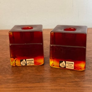 May include: Two amber glass candle holders with a square shape and a small round depression in the top. The bottom of each holder has a label that reads "Lindshammar Sweden Orrefors".