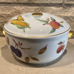 May include: White ceramic casserole dish with a lid, featuring a gold trim and a floral design with corn, peaches, and plums.
