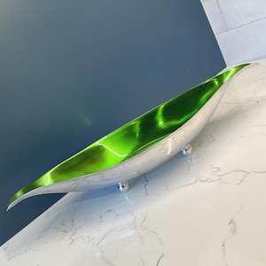 May include: A long, silver-colored decorative bowl with a vibrant green interior. The bowl has a sleek, elongated shape and rests on three small, round silver feet. The green interior has a marbled effect.
