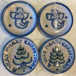 May include: Four ceramic plates with a blue rim. Two plates feature a blue angel design with stars, and the other two have a blue Christmas tree with the words "A Very Merry Christmas" in blue around the tree.