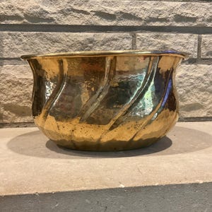 May include: A polished brass bowl featuring a wide, flared rim and a series of diagonal, recessed fluted channels along the exterior surface. The metal has a reflective, golden finish with a subtle hammered texture throughout the body.