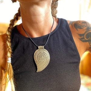 Può includere: Una donna indossa una canotta nera con una collana con un ciondolo d'argento a forma di lacrima con dettagli. Il ciondolo è appeso a un cordoncino nero.