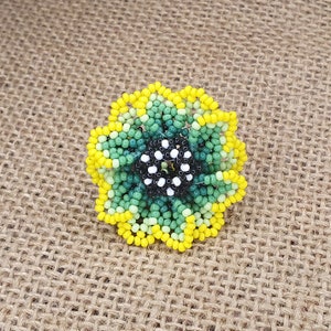 Peut inclure: Une fleur en perles avec un centre vert et jaune et un centre noir et blanc. La fleur est faite de petites perles et est un artisanat fait main.
