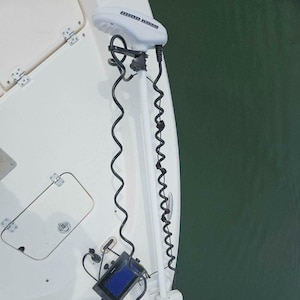 May include: A white electric trolling motor with a black coiled cord attached to a boat. The motor is mounted on the back of the boat and is in the water. A small black and white fish finder is mounted on the boat's deck.