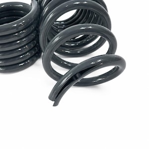 May include: Two dark grey metal coil springs with a smooth, glossy finish. The springs feature a cylindrical shape with multiple tight loops and a distinct open end, showing the hollow interior structure of the thick, durable metal wire construction.