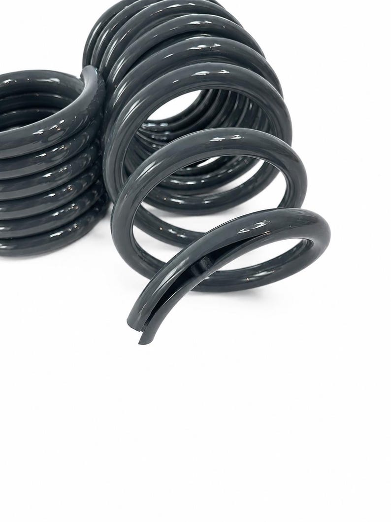 May include: Two dark grey metal coil springs with a smooth, glossy finish. The springs feature a cylindrical shape with multiple tight loops and a distinct open end, showing the hollow interior structure of the thick, durable metal wire construction.