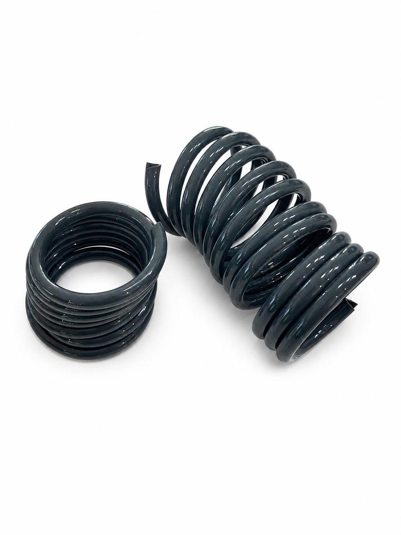 May include: Two dark grey, glossy, coiled metal springs. One spring is positioned vertically in a tight, cylindrical spiral shape, while the other is laid horizontally, showing a circular opening through the centre of the coiled wire structure.