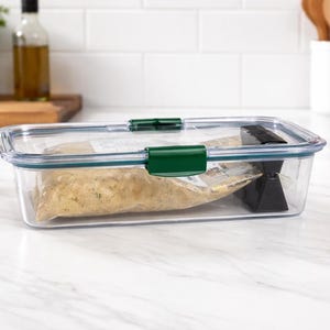 May include: A clear rectangular food storage container with a green snap-on lid. The container holds a sealed bag of food and a black object. The container is on a white countertop.