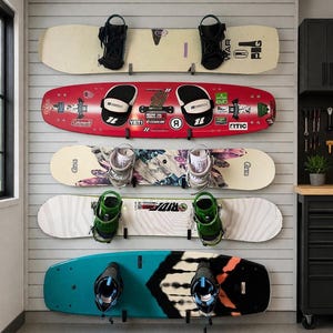 Puede incluir: Cinco tablas de snowboard de varios colores y diseños están montadas en una pared blanca. Las tablas de snowboard tienen fijaciones negras y están listas para usar. Las tablas son rojas, blancas, turquesas y con pegatinas.