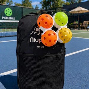 Op de afbeelding: Zwarte rugzak met een mesh-vak, met vier pickleballs in oranje, groen en geel. De ballen worden in een witte houder gehouden. De rugzak heeft de merknaam "niup" en het model "MX-23" erop gedrukt.