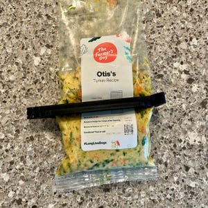 以下が含まれることがあります： The Farmer's Dogの「Otis's Turkey Recipe」と書かれた、透明な犬用フードの袋。黒いクリップで密封されています。食べ物には野菜と七面鳥が含まれています。「#LongLiveDogs」の文字が見えます。