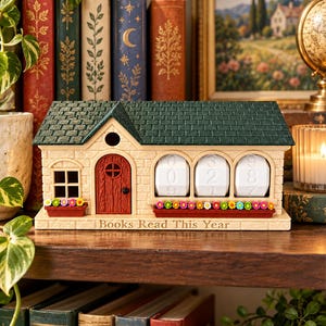 May include: A house-shaped book tracker with a dark green roof, beige brick walls, and a red door. It features three white rotating number dials and two window boxes filled with colorful flowers. The base is inscribed with "Books Read This Year."