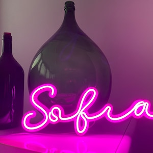 Può includere: Un cartello al neon rosa con il nome "Sofia" scritto in corsivo. Il cartello è appoggiato su un tavolo davanti a una grande bottiglia di vetro.