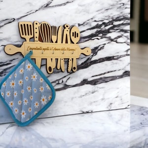Op de afbeelding: Houten keukenutensilienhouder in de vorm van een deegroller met de tekst "L'ingrediente segreto è l'Amore della Mamma!" De houder heeft uitsparingen voor keukengerei en haken om op te hangen. Een blauwe en witte ovenwant met een madeliefjespatroon hangt aan de houder.