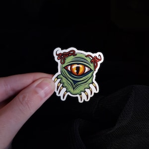 Puede incluir: Pegatina de monstruo verde con un solo ojo naranja grande y tentáculos rojos. El monstruo tiene garras afiladas y está sobre un fondo blanco.