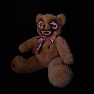 Puede incluir: Un oso de peluche marrón con una corbata de lazo roja y una boca cosida. Los ojos del oso están cosidos y hay manchas de sangre en su pelaje.