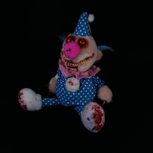 May include: A disturbing clown doll with a blue polka-dot outfit, a pink nose, and a matching hat. The doll has red eyes, a stitched mouth with visible teeth, and blood-like stains on its body and feet. The doll is sitting against a black background.