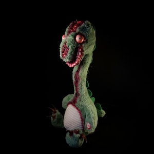 May include: A green plush dinosaur toy with a zombie theme. The toy has exposed red flesh and a stitched appearance. It features large pink eyes and a long neck, with a striped belly and clawed feet. The background is black.
