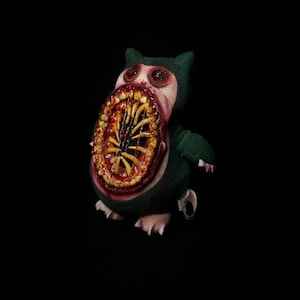 May include: A plush toy in the shape of a green creature with a large, open mouth revealing rows of yellow teeth and a bloody interior. The toy has small arms, feet, and red eyes, set against a black background.