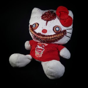 Puede incluir: Un peluche blanco con un lazo rojo y una camiseta roja. El juguete tiene una apariencia grotesca de zombi, con dientes expuestos, un cerebro y detalles sangrientos. La camiseta tiene un estampado gráfico.