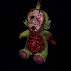 Puede incluir: Un juguete de peluche verde con el pecho abierto y un esqueleto sangriento visible. El juguete tiene una cara grotesca y sangrienta con un solo ojo que sobresale.