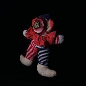 May include: A handmade doll with a large, realistic eyeball for a head. The doll has a red ruffled collar, striped red and blue legs, and white socks. The doll's hat is blue. The background is black.