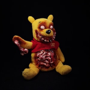 Pode incluir: Um urso de peluche amarelo com uma camisola vermelha com o texto Winnie the Pooh. O urso apresenta olhos brancos, uma boca larga cheia de dentes e órgãos internos expostos e manchados de sangue no tronco e num braço.