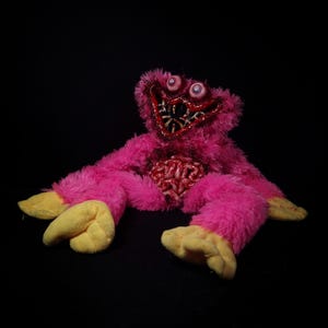 Può includere: Un peluche con pelliccia rosa brillante, mani e piedi gialli e una bocca larga e scura piena di denti appuntiti. La creatura ha due occhi sporgenti e una struttura dettagliata simile a un cervello esposta nella zona del torace.
