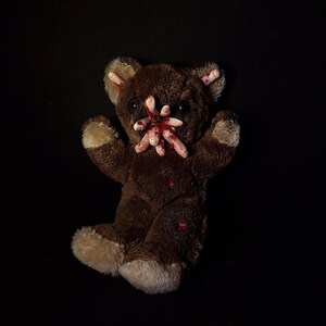 Puede incluir: Un oso de peluche marrón con detalles en beige en las patas y orejas. Varios apéndices pequeños y pálidos con forma de dedos emergen de su boca. Manchas rojas son visibles en el pelaje, sugiriendo un tema de terror.